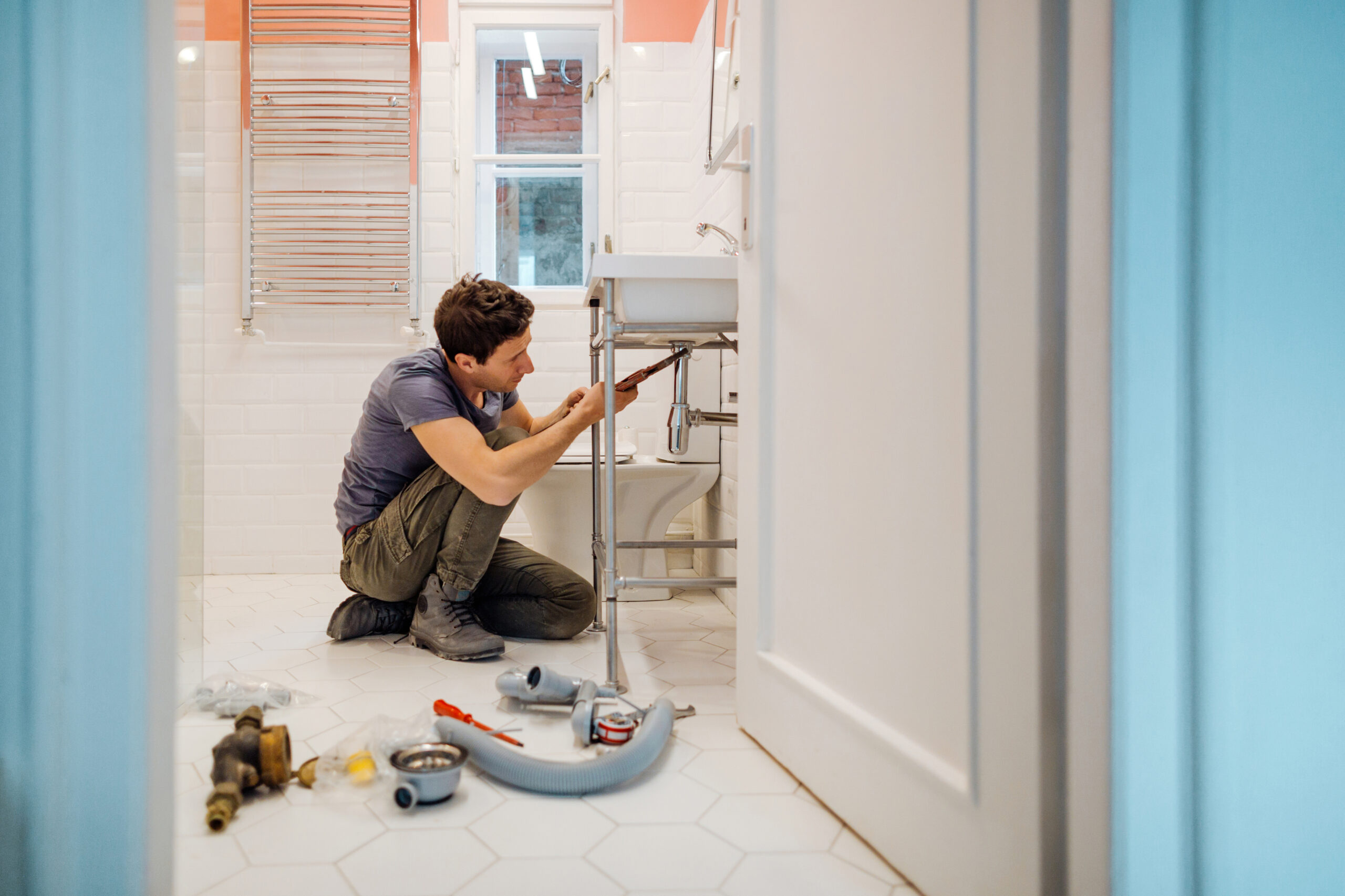 an image about In a tiled bathroom, a person is kneeling on the floor, working diligently on the plumbing under a sink. Various tools and plumbing parts are scattered around, marking the location as an impromptu workspace.