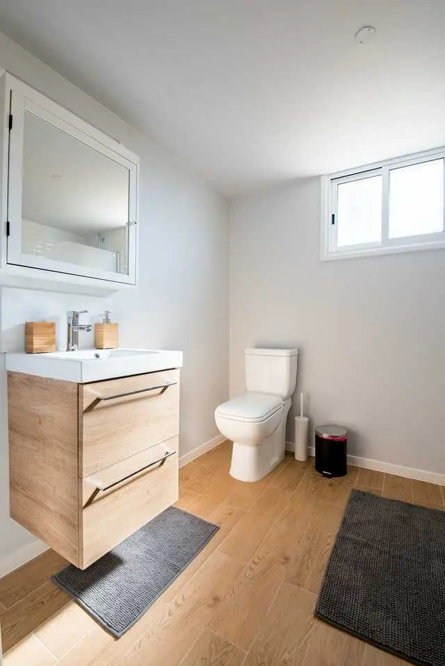 an image about A modern bathroom with wooden vanity, white toilet, and a small window.