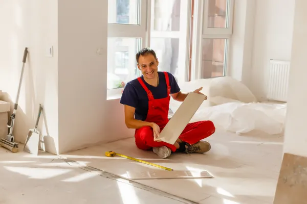 an image about A man in overalls sitting on the floor holding a piece of floor tiles material contemplating tile installation.